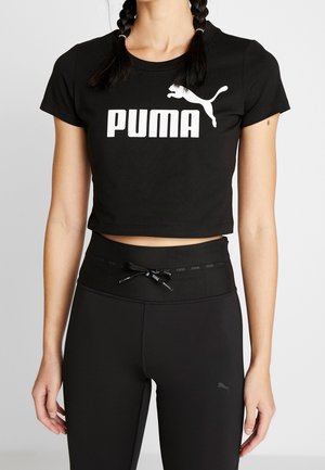 Kvinde iført sort, kort Puma t-shirt og sort, høj talje leggings med snor mod ensfarvet baggrund.