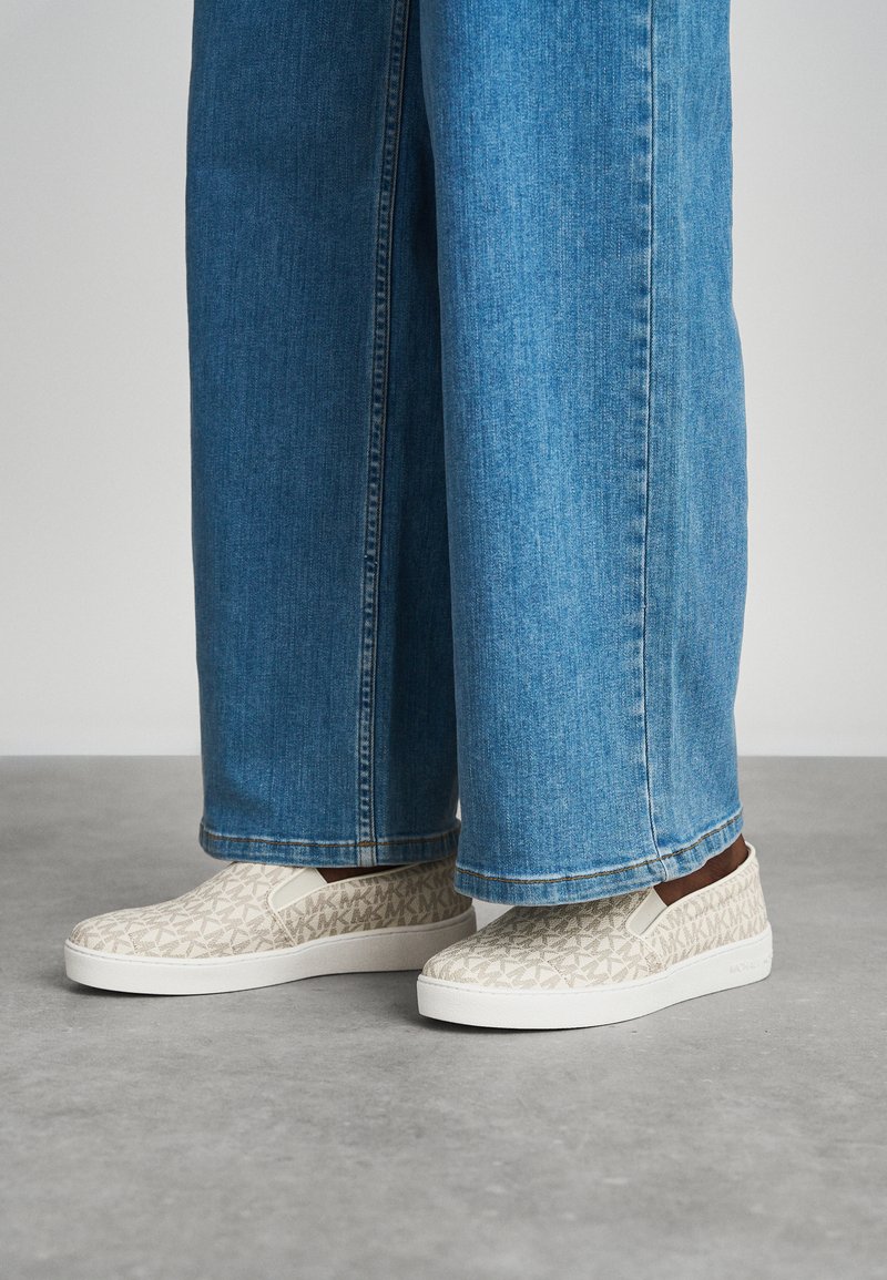 Slip-on shoes with a textured beige fabric featuring a geometric pattern and white rubber sole, paired with wide blue denim pants.