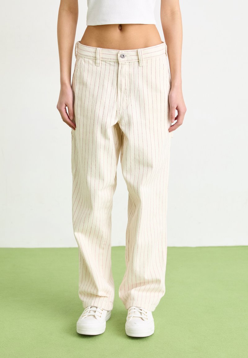 Cream-colored, wide-leg pants with red vertical stripes and a button closure. Worn with white sneakers. Smooth fabric, slightly textured.