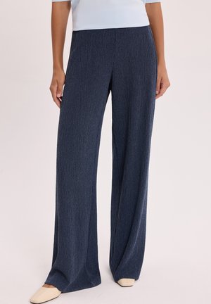 Kobieta w granatowych, prążkowanych szerokich spodniach i kremowych butach na kwadratowym nosku stojąca na jednolitym tle.