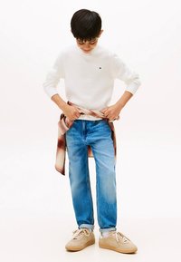 Weißes Sweatshirt, hellblaue Jeans, beige Sneaker. Ein kariertes Flanellhemd, um die Taille gebunden, sorgt für Farbe und Textur.