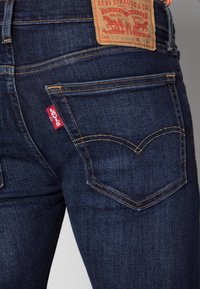 Mörkblå denimjeans med närbild på bakfickorna. Har gul söm och en röd etikett på vänster ficka. Lädermärke med varumärket synligt.