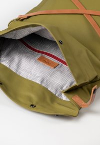 Borsa verde oliva con un esterno texturizzato, dotata di una tracolla in pelle marrone e un interno a motivo a griglia. Include una tasca con zip rossa.