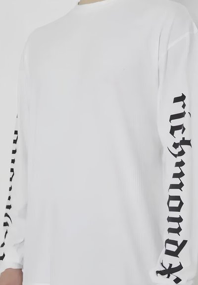 Camicia a maniche lunghe bianca con testo nero lungo le maniche. Il tessuto sembra liscio con una vestibilità rilassata e un design a girocollo.