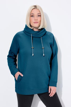 Femme avec une coupe au carré blonde, portant un sweat à capuche sarcelle avec un col montant et des cordons rayés, debout avec une main dans la poche.