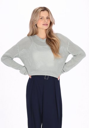 Lysegrøn strikket sweater med V-hals og løs pasform, kombineret med marineblå bukser med vidde. Strukturen er åbenstrikket, hvilket tilfører visuelt interesse.
