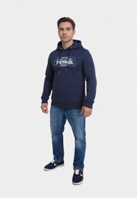 Námornícka mikina s grafickým logom "PETROL", s kapucňou na sťahovanie a rebrovanými manžetami, v kombinácii s modrými džínsami a námorníckymi teniskami.