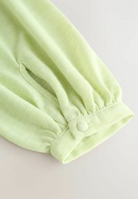 Poignet de manche longue vert clair avec élastique froncé et fermeture par bouton recouvert de tissu sur un tissu doux et texturé.