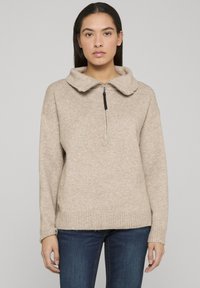 Pull beige zippé avec un grand col, en tissu tricoté texturé. Ourlet et poignets côtelés. Associé à un jean bleu foncé.