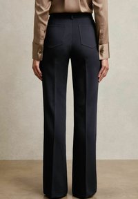 Marineblaue Schlaghosen mit zwei Gesäßtaschen, glatter Stoffoberfläche und einem figurbetonten Bund. Kombiniert mit einer hellbraunen Langarmbluse.