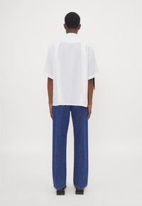 Camicia bianca a maniche corte con vestibilità rilassata, abbinata a jeans blu larghi in denim. La camicia ha un orlo dritto e un colletto semplice.