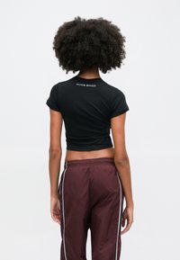 Crna skraćena majica s kratkim rukavima, s bijelim "NIKE SHOX" logotipom na leđima, u kombinaciji s maroon treniranim hlačama s bijelim bočnim trakama.