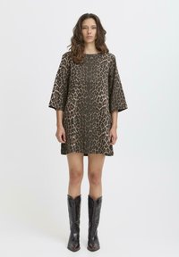 Leopardenmuster-Kleid in Braun und Schwarz, mit lockerem Schnitt, weiten Ärmeln und schneidet bis zur Mitte des Oberschenkels. Kombiniert mit schwarzen Cowboystiefeln.
