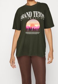 Camiseta de color verde oliva con un gráfico que presenta una escena montañosa, un atardecer y el texto que dice "PARQUE NACIONAL GRAND TETON". Mangas cortas, corte relajado.