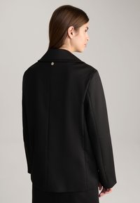 Zwarte blazer van gladde stof, voorzien van een geknipte kraag, gestructureerde schouders en een enkele decoratieve knoop op de rug.