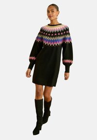 Rochie tricotată neagră cu un guler cu model în nuanțe de roz, albastru și verde. Are mâneci tip ballon și este asortată cu cizme negre până la genunchi.