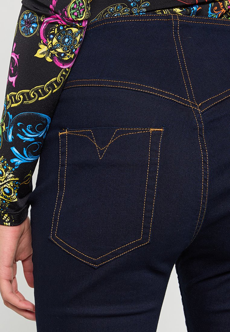 Osoba w ciemnoniebieskich dżinsach z pomarańczowym przeszyciem i kolorowej, wzorzystej bluzie z długim rękawem, z ręką opartą na boku.