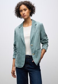 Teal corduroy bleiseri, jossa on kaksi nappia, kauluksessa lovi ja etutaskut, käytössä valkoisen paidan päällä ja yhdistetty tummiin farkkuihin.