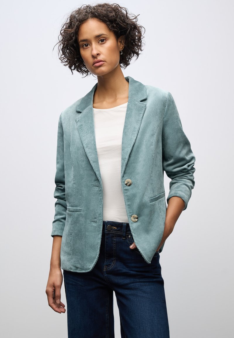 Teal corduroy bleiseri, jossa on kaksi nappia, kauluksessa lovi ja etutaskut, käytössä valkoisen paidan päällä ja yhdistetty tummiin farkkuihin.