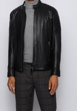 Ein Mann trägt eine schwarze gesteppt Lederjacke über einem grauen Rollkragenpullover und grauen karierten Hosen vor einem schlichten grauen Hintergrund.