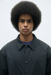 Svart jacka med krage och knappstängning, bärs över en blå skjorta. Modellen har en rund, voluminös afrofrisyr.