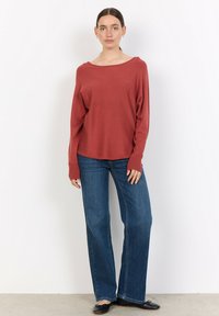 Rostfärgad långärmad topp med bred ringning, kombinerad med blå utsvängda jeans. Outfiten har en avslappnad passform och ribbat mönster.