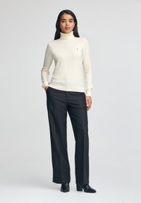 Maglione a collo alto crema con maniche lunghe, abbinato a pantaloni neri su misura. Accento del logo ben visibile sul petto sinistro. Texture liscia.