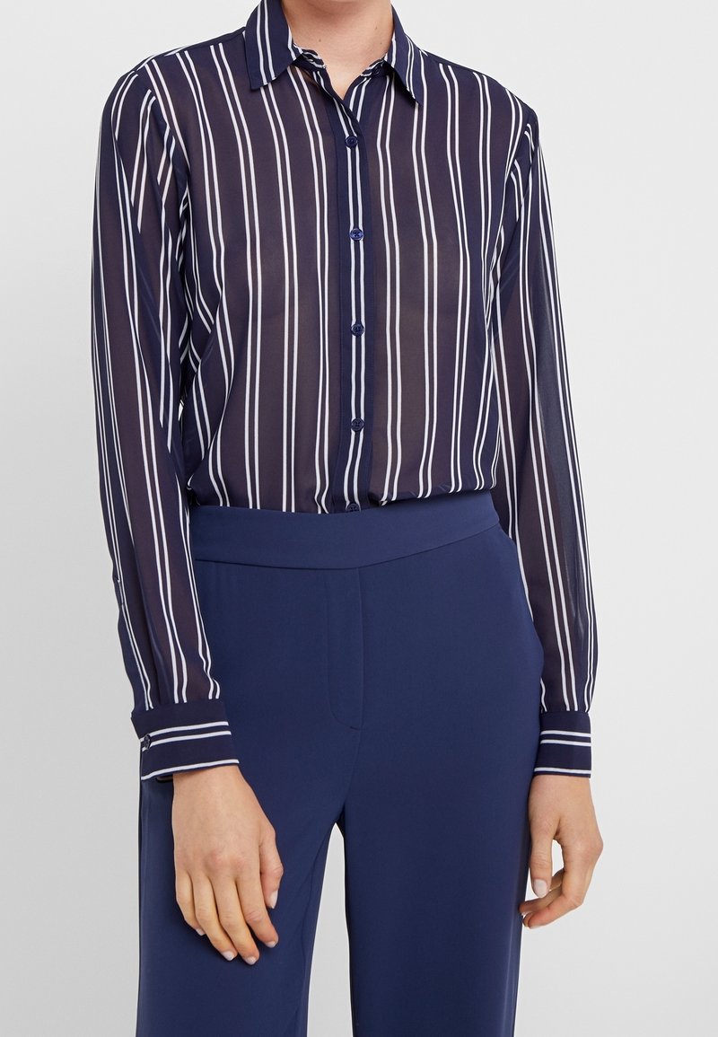 Femme portant une chemise longue bleu marine transparente à rayures verticales blanches et un pantalon bleu marine taille haute assorti.