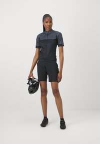 Maillot de cyclisme à manches courtes gris foncé avec une fermeture éclair, associé à un short noir et des chaussures de cyclisme, tenant un casque noir. Design élégant et sportif.