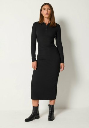 Femme debout contre un mur blanc, portant une longue robe noire cintrée avec des boutons et des bottines noires.