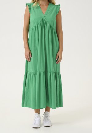 Femme portant une robe midi verte sans manches avec des épaules à volants et des baskets blanches, debout devant un fond uni.