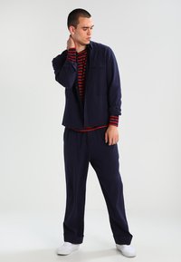 Giacca blu navy sopra una maglietta a strisce rosse e blu navy, abbinata a pantaloni blu navy. Il modello sta in piedi con una mano sul collo, indossando scarpe bianche.