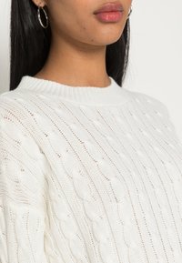 Weißer Strickpullover mit strukturiertem Kabelmuster und geripptem Halsausschnitt. Nahaufnahme der komplizierten Details des Stoffes.