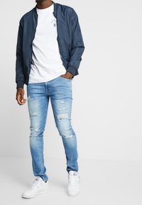 Marinblå bomberjacka, vit grafisk T-shirt, ljusblå slitna jeans och vita sneakers. Avslappnad outfit med olika texturer och färger.