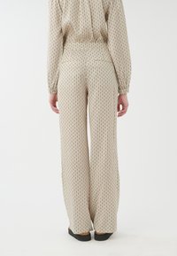 Pantalon large beige à motifs, avec un design en losanges répété, doté d'une taille haute et d'une ceinture élastique, associé à des chaussures noires.