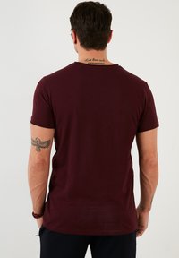Burgundska majica s kratkimi rokavi s klasičnim okroglim ovratnikom, izdelana iz mehke tkanine. Ponuja opazno tetovažo na desni roki.