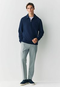 Giovane uomo in piedi contro un muro bianco, indossando un pullover navy a zip corta, pantaloni grigio chiaro e sneakers navy, con una mano in tasca.