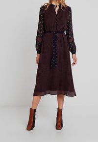  långärmad klänning i knälång modell med mörk blommönstrad tyg, med knytdetalj vid halsen och cinchad midja. Matchas med tvåfärgade ankelstövlar.
