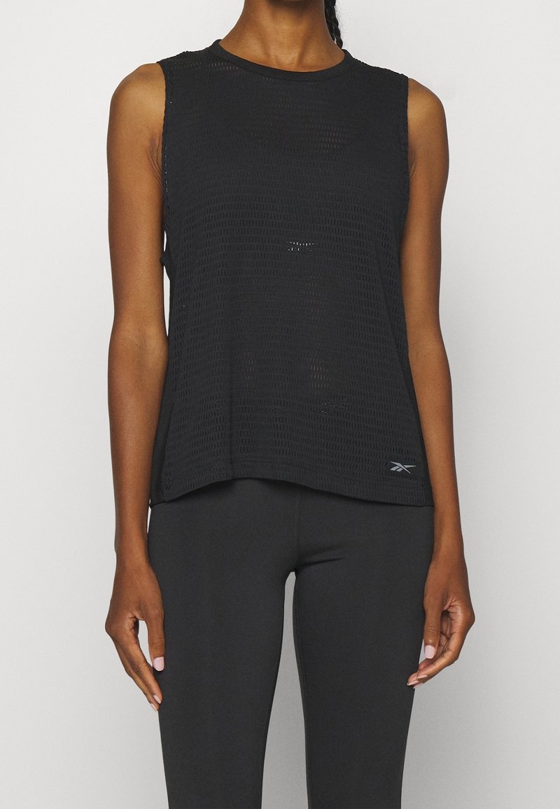 Personne portant un haut en maille noir sans manches et un legging noir, debout devant un fond clair uni.