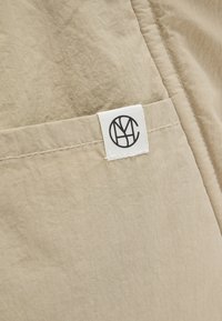 Μπεζ ύφασμα με ραμμένη ραφή και μικρό λευκό ορθογώνιο ετικέτα που φέρει κυκλικό μαύρο λογότυπο με στυλιζαρισμένα επικαλυπτόμενα γράμματα.