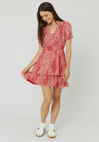 Robe rouge à manches courtes avec un motif floral, ceinture à la taille et jupe à volants superposés. Des baskets blanches et des chaussettes de cheville complètent le look.