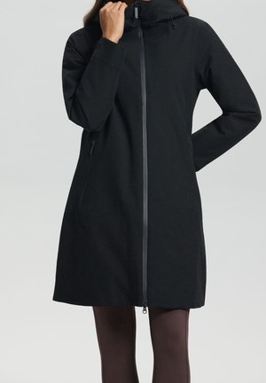 Femme portant un manteau long noir ajusté avec une fermeture éclair à l'avant, un col haut et des poches latérales, debout devant un fond gris uni.
