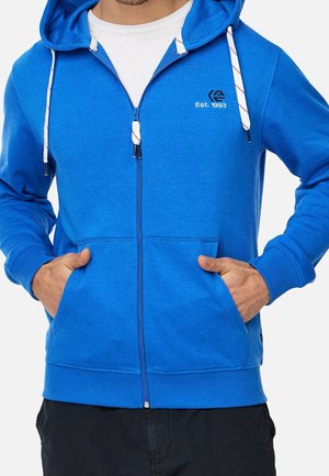 Homme portant un sweat à capuche bleu avec fermeture éclair, cordons blancs et texte « Est. 1993 » sur la poitrine, les mains dans les poches avant.