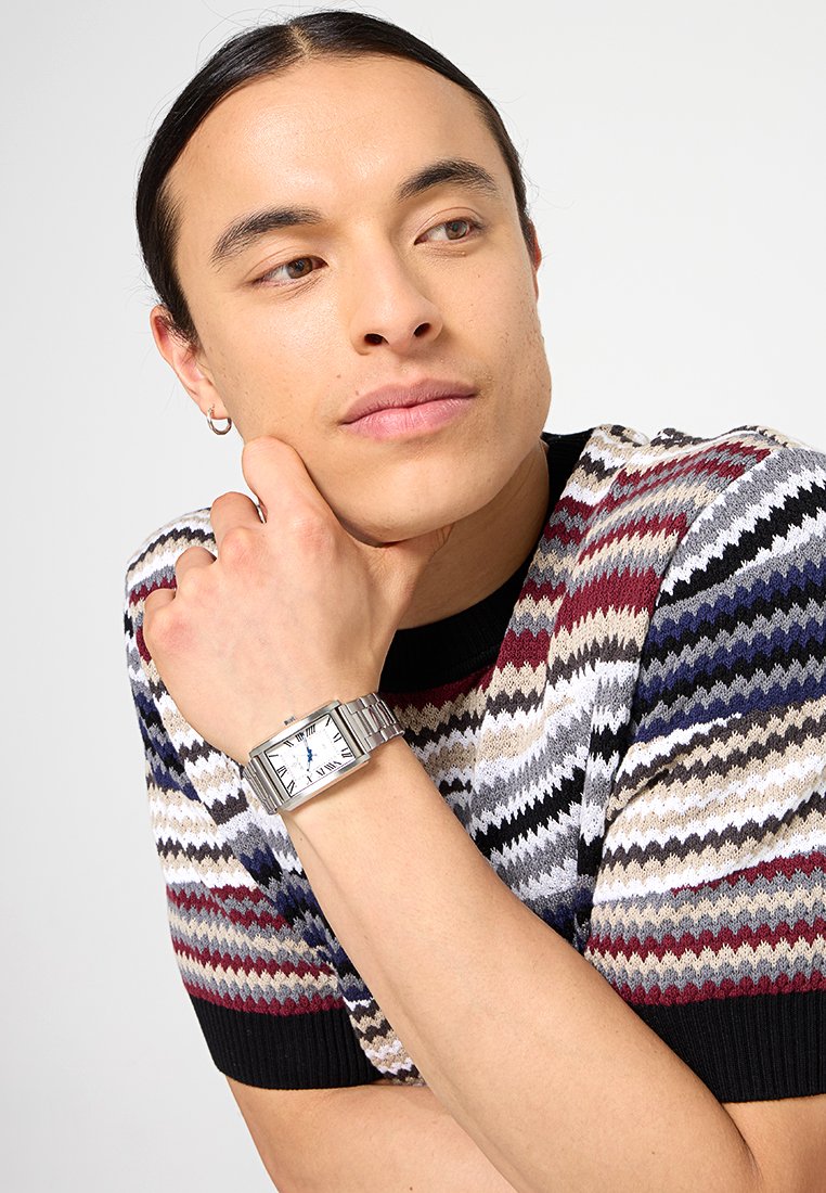 Montre en métal argenté avec un cadran rectangulaire, des chiffres romains et un bracelet texturé, portée sur un poignet avec un pull à rayures en zigzag multicolores.