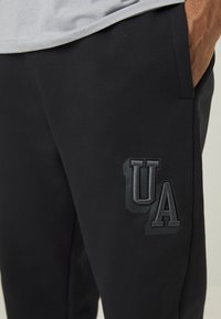 Zwarte trainingsbroek van zacht materiaal, met een geplayeerde donkere "UA"-logo op de linker bovenbeen en zijzakken voor extra functionaliteit.