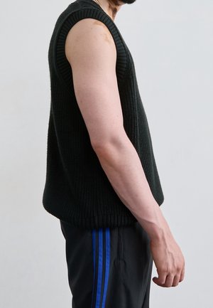 Mann trägt ärmellosen schwarzen Strickpullover und schwarze Hose mit blauen Streifen, steht vor schlichtem hellem Hintergrund.