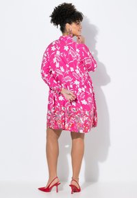 Helles pinkes Blumenkleid mit langen Ärmeln, das weiße Blumenmuster und einen ausgestellten Saum aufweist. Kombiniert mit roten High-Heels mit Slingback.