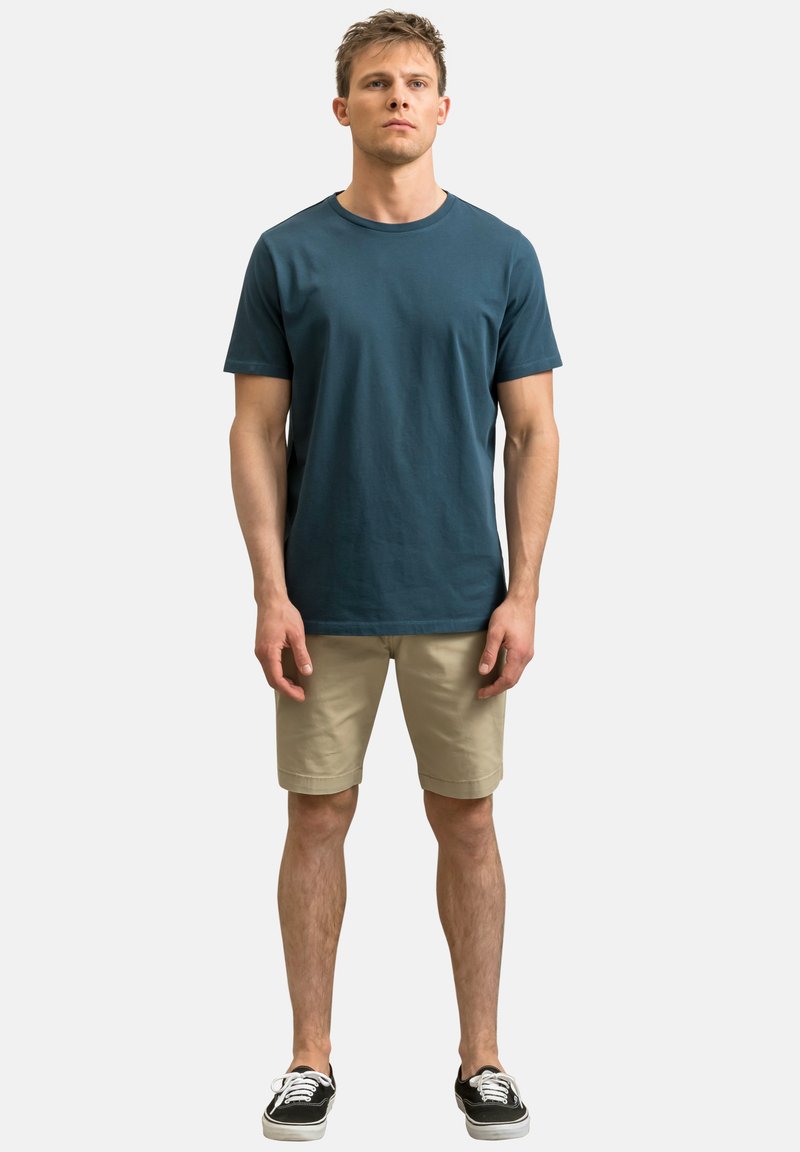 Heren teal katoenen T-shirt met korte mouwen, gecombineerd met beige shorts en zwarte sneakers. Eenvoudig ontwerp, zachte textuur en normale pasvorm.