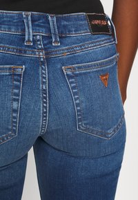 Jeans de mezclilla azul con un diseño ajustado, costuras en contraste de color naranja, dos bolsillos traseros y un parche de cuero negro con marca en la cinturilla.