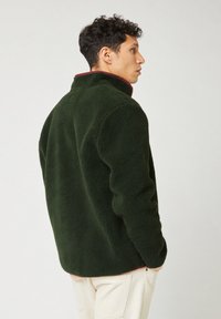 Giacca in pile verde con superficie texturizzata, collo alto e foderatura interna bordeaux a contrasto. Presenta una vestibilità comoda e orlo arrotondato.